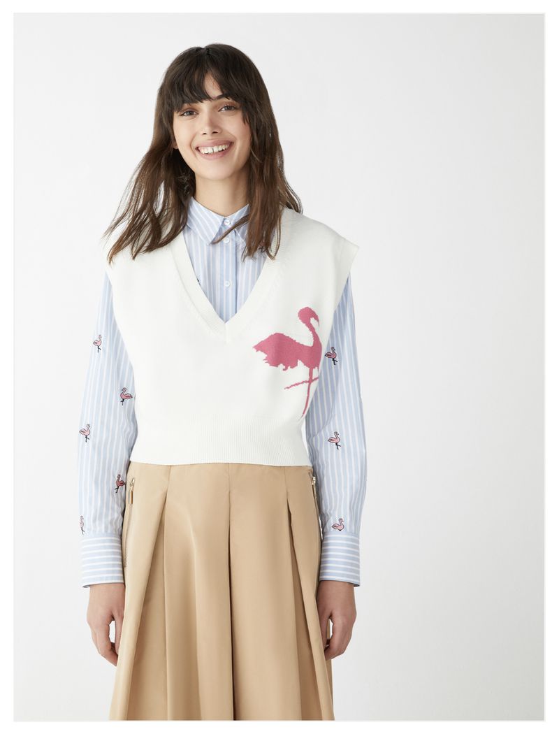 A woman smiles wearing a sweater vest with a pink flamingo design over a striped shirt and khaki skirt against a white backdrop.
