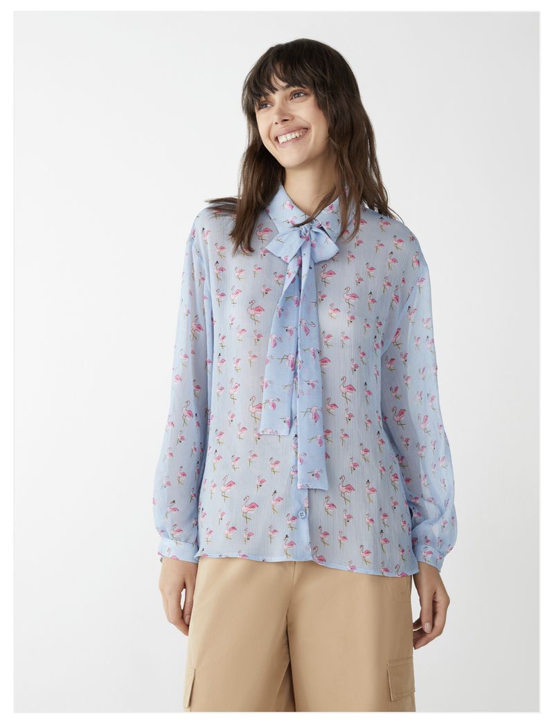 Smiling woman in a light blue long-sleeve flamingo-print shirt with a tie, paired with khaki cargo shorts, isolated on white.