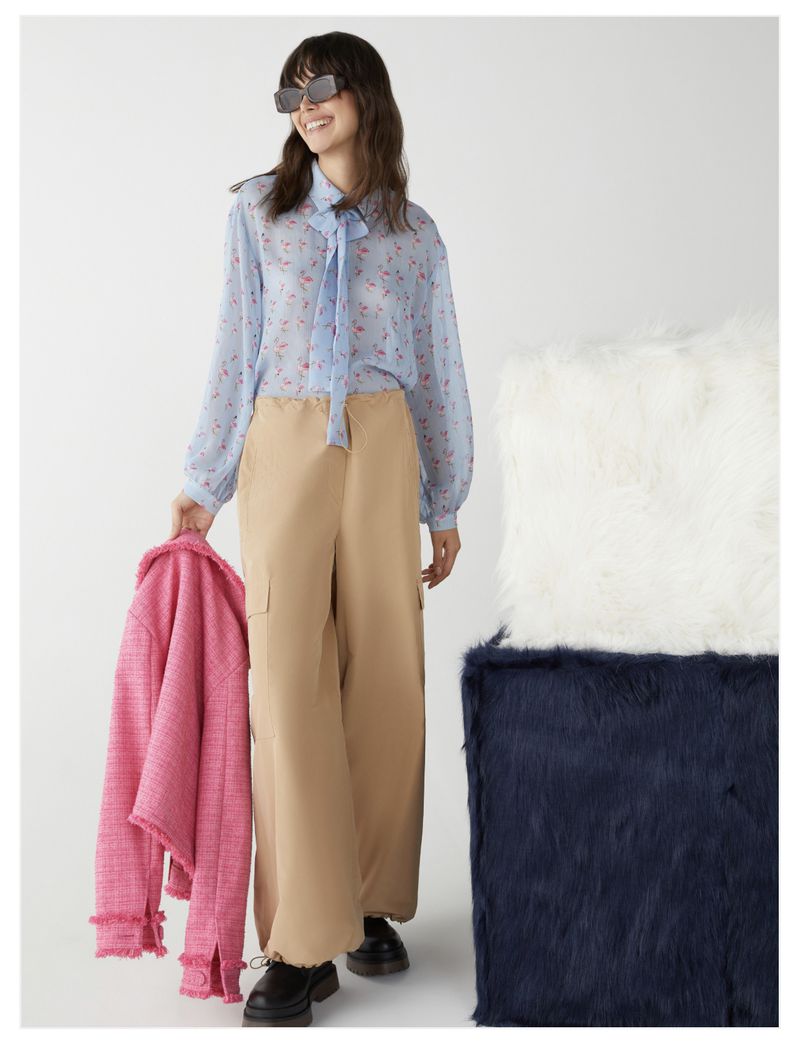 Woman in floral blouse and beige pants holds pink jacket, posing in a studio with textured white and blue boxes in background.
