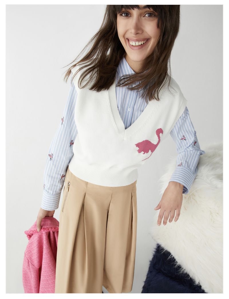 A woman smiles in a studio, wearing a striped shirt, sweater vest with a bird design, and tan pants, while holding a pink jacket.
