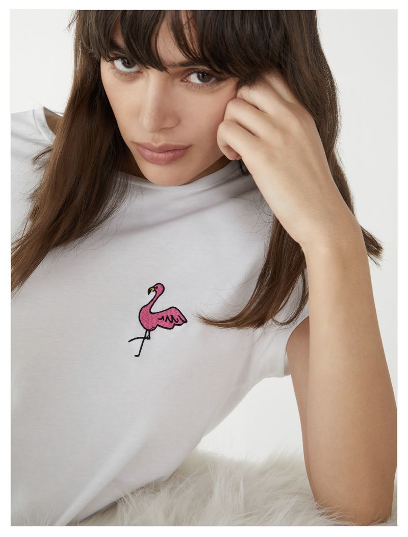 Brunette woman with hand near face wearing a white t-shirt with a pink flamingo embroidered on the front, posing indoors.