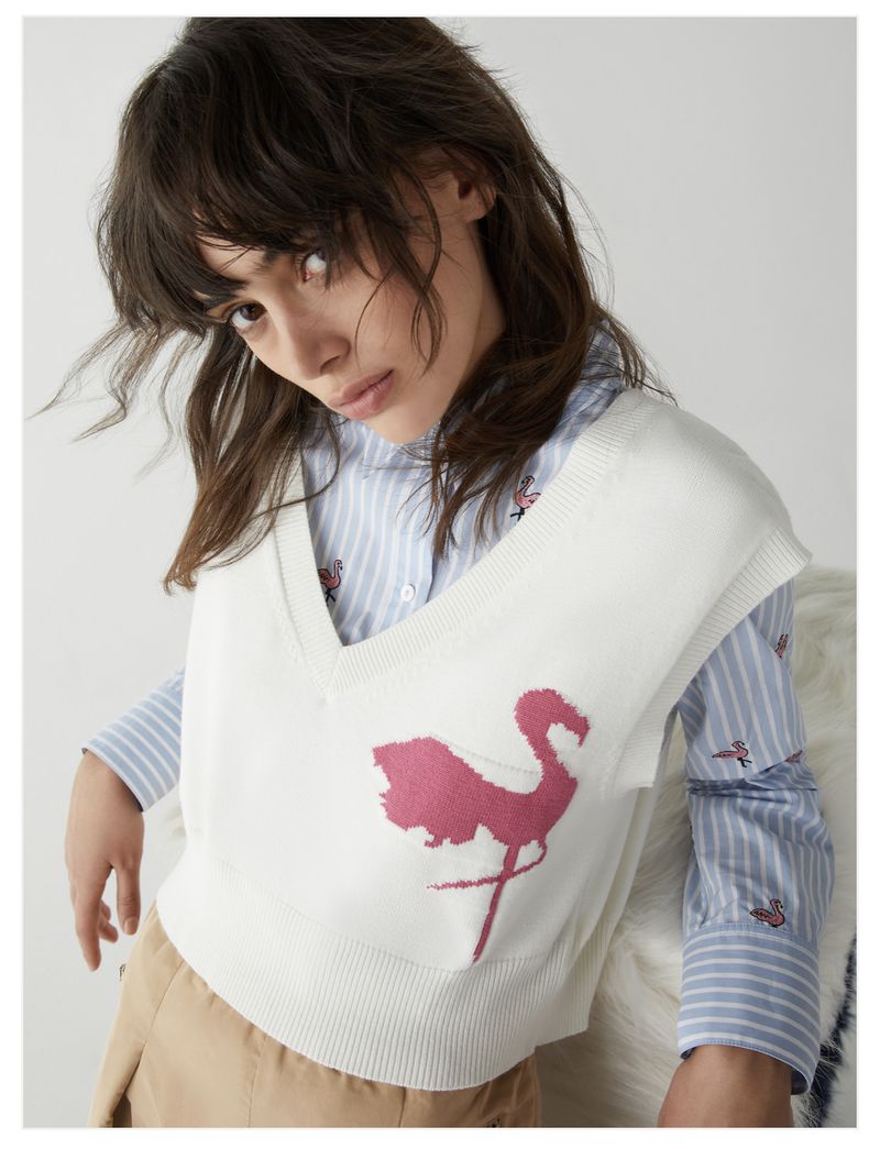 A fair-skinned young woman wears a striped shirt and flamingo-print knit sweater-vest indoors against a neutral background in a studio shot.