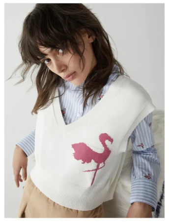 A fair-skinned young woman wears a striped shirt and flamingo-print knit sweater-vest indoors against a neutral background in a studio shot.