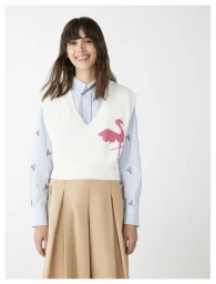 Model Rosa Kirakosyan smiles wearing a white flamingo knit vest over a blue striped shirt and beige trousers against a white background.
