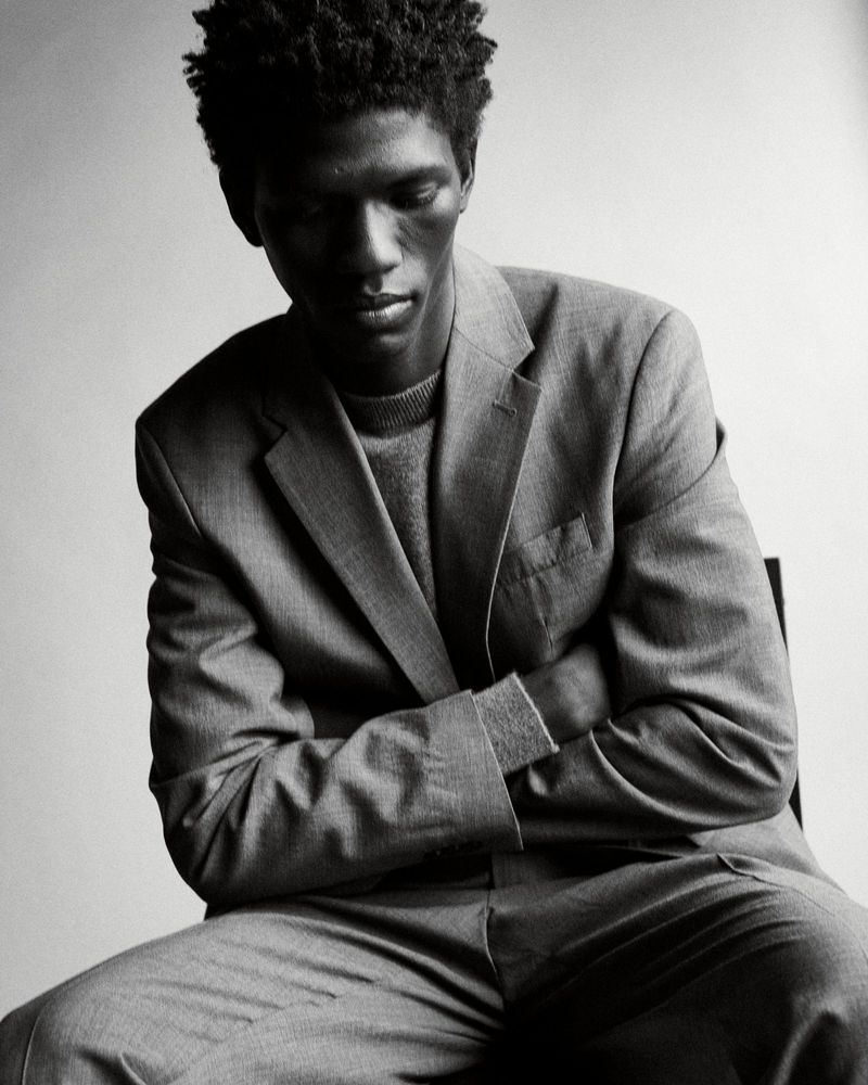 Monochrome portrait of a man in a grey suit jacket and sweater, seated with arms crossed, in a studio setting with soft lighting.