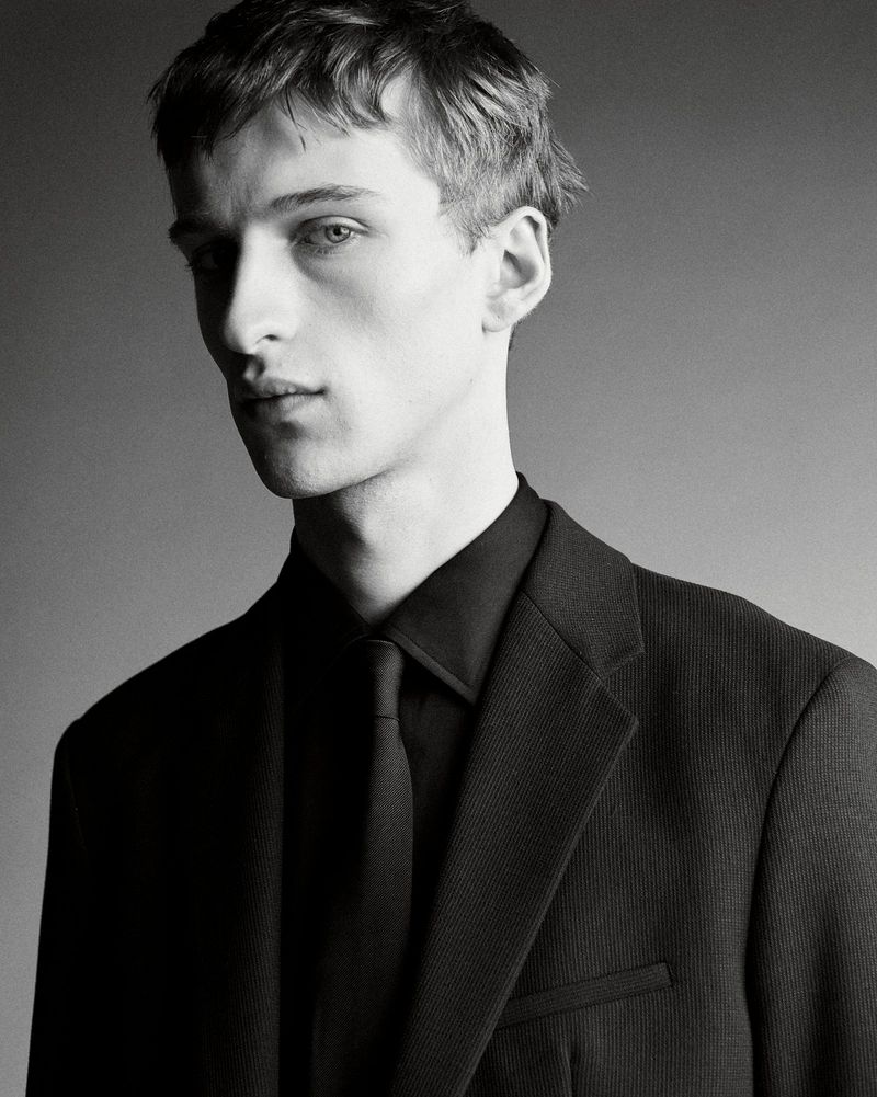 Portrait of a young male model wearing a black suit jacket, black shirt, and matching black tie against a grey background.