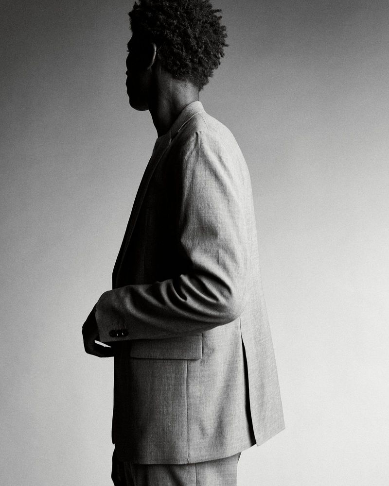 Black man wearing a tailored suit jacket in profile view against a neutral gradient studio background