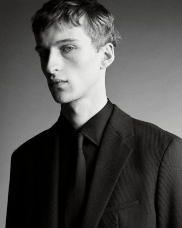 Portrait of a young male model wearing a black suit jacket, black shirt, and matching black tie against a grey background.