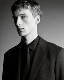 Portrait of a young male model wearing a black suit jacket, black shirt, and matching black tie against a grey background.