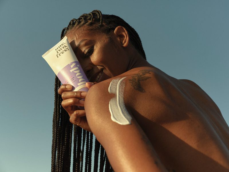 Woman applies body lotion to shoulder outdoors in natural sunlight against a blue sky, showcasing beauty and skincare routine.