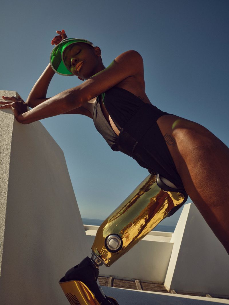 A woman with a prosthetic leg poses confidently in a black swimsuit and green visor against a blue sky and bright white architecture.