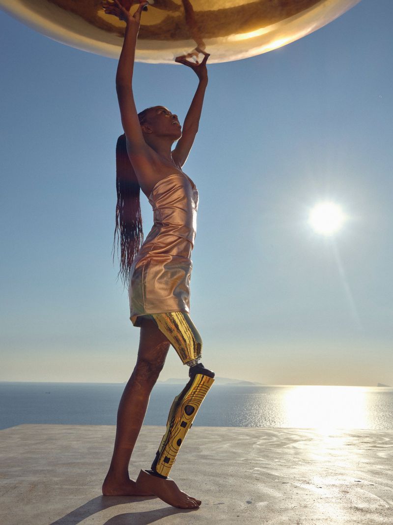 Woman with prosthetic leg in gold dress holds a large, gold, reflective sphere outdoors against a background of blue sky and ocean.