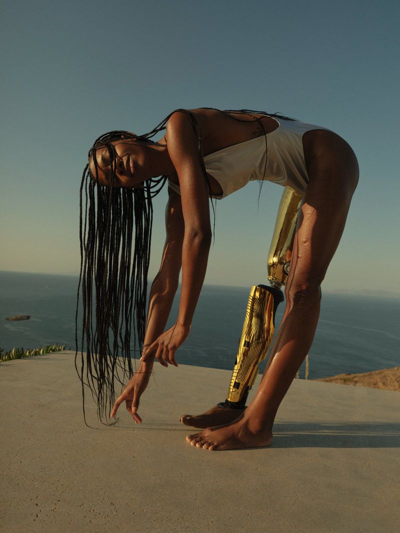 Athletic woman with a prosthetic leg bends over on an outdoor terrace against a blue sky and ocean, showcasing strength and resilience.