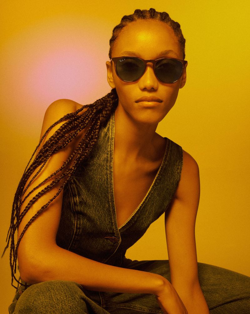 Portrait of a young woman wearing sunglasses and a denim vest sitting against a solid yellow background in a studio environment.