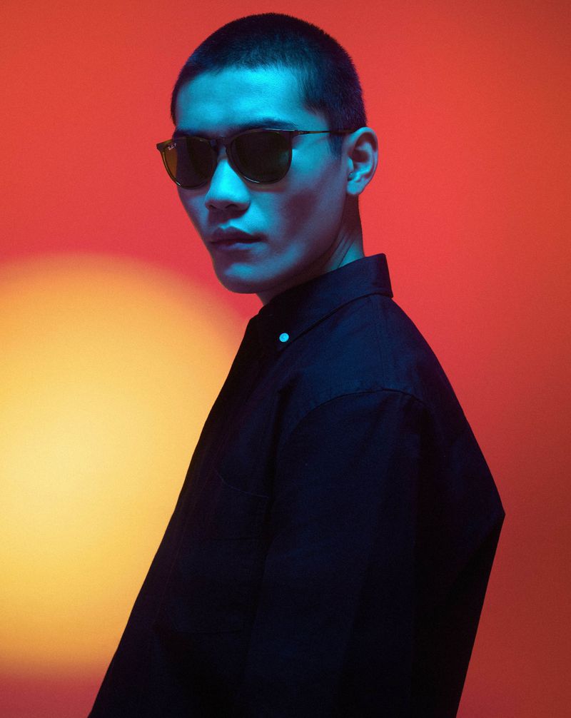 An Asian man in a black shirt and sunglasses is brightly illuminated with blue light against an orange gradient background in a studio shot.