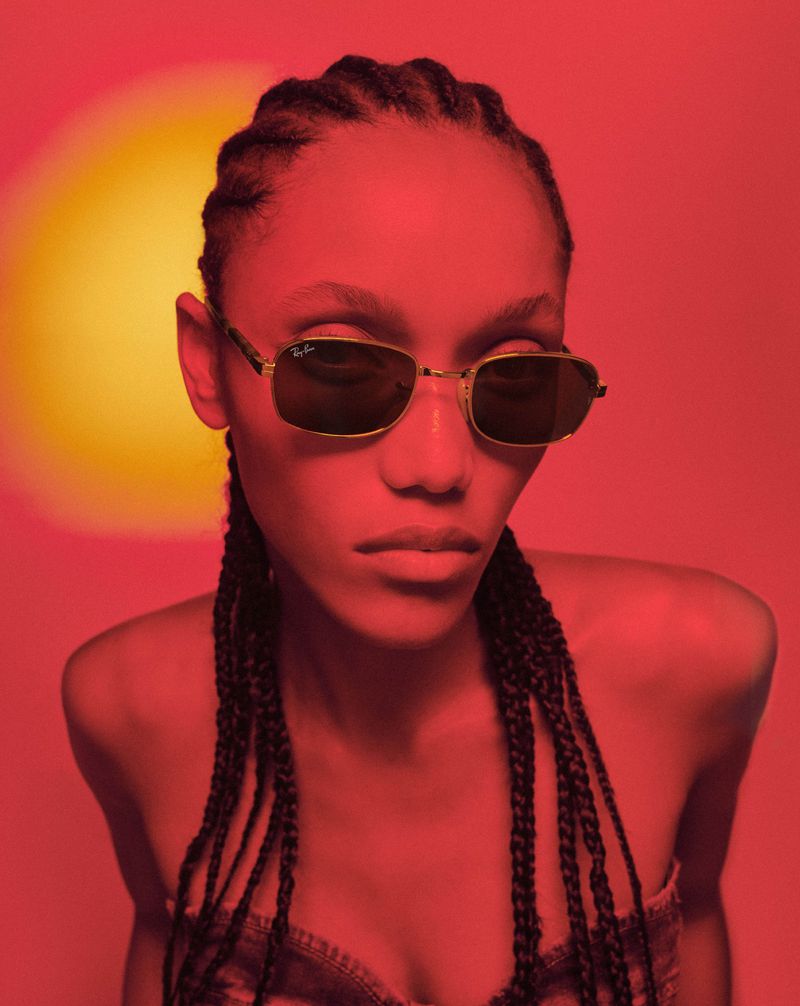 Close-up of an adult female with braids wearing sunglasses, captured in a photo studio against a solid red background with a spotlight effect.
