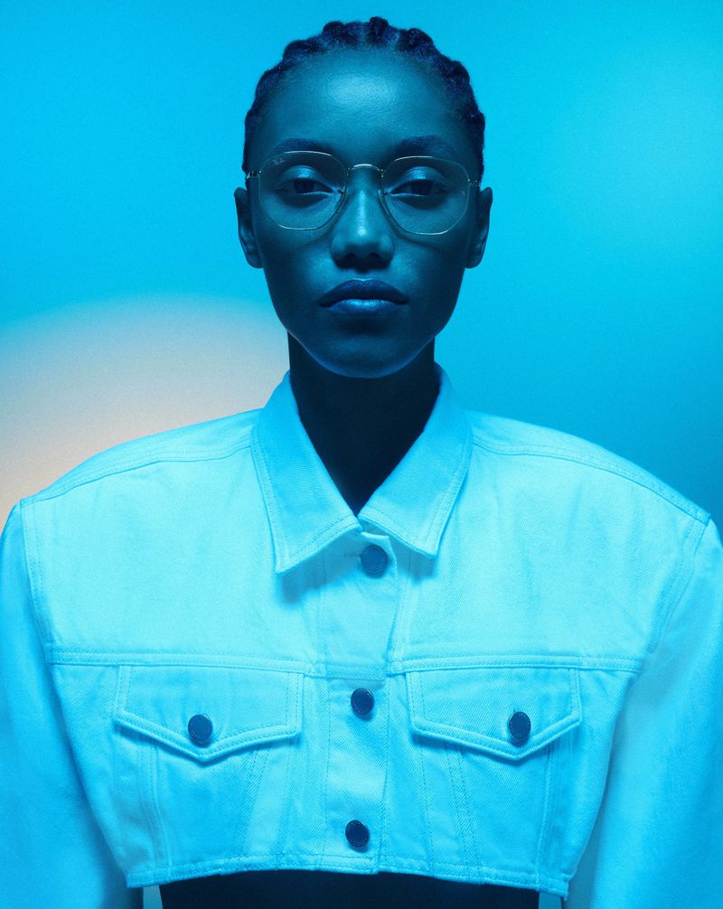 Close-up of an adult woman with eyeglasses in a white jean jacket against a blue background in a portrait-style studio shot.