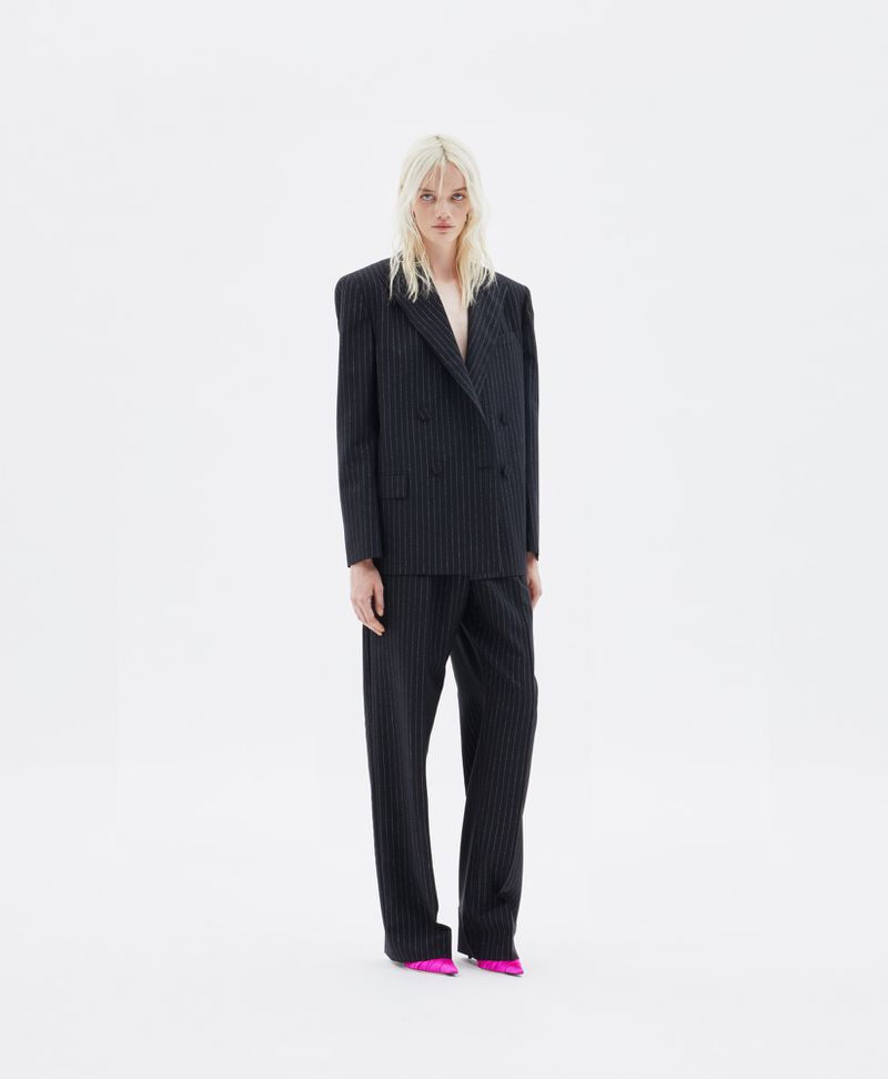 A fashion portrait showcases an adult female in a dark pinstripe suit and pink heels set against a bright white backdrop in a photo studio.