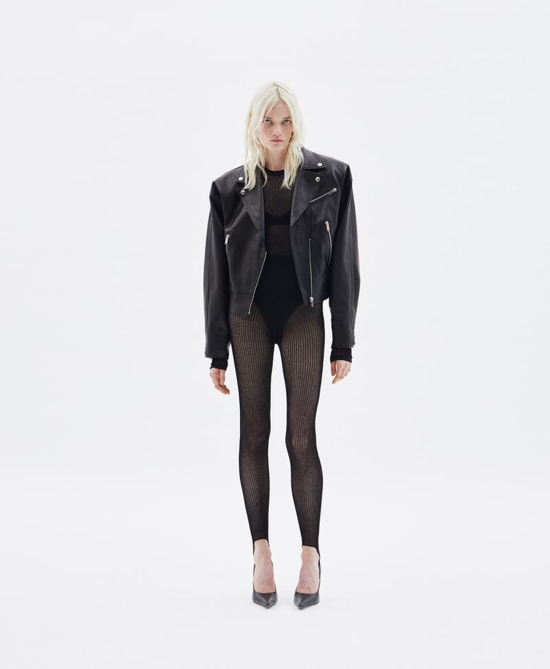 Full-length studio shot of an adult female model with platinum blonde hair wearing a black leather jacket, bodysuit, fishnet stockings, and high heels.