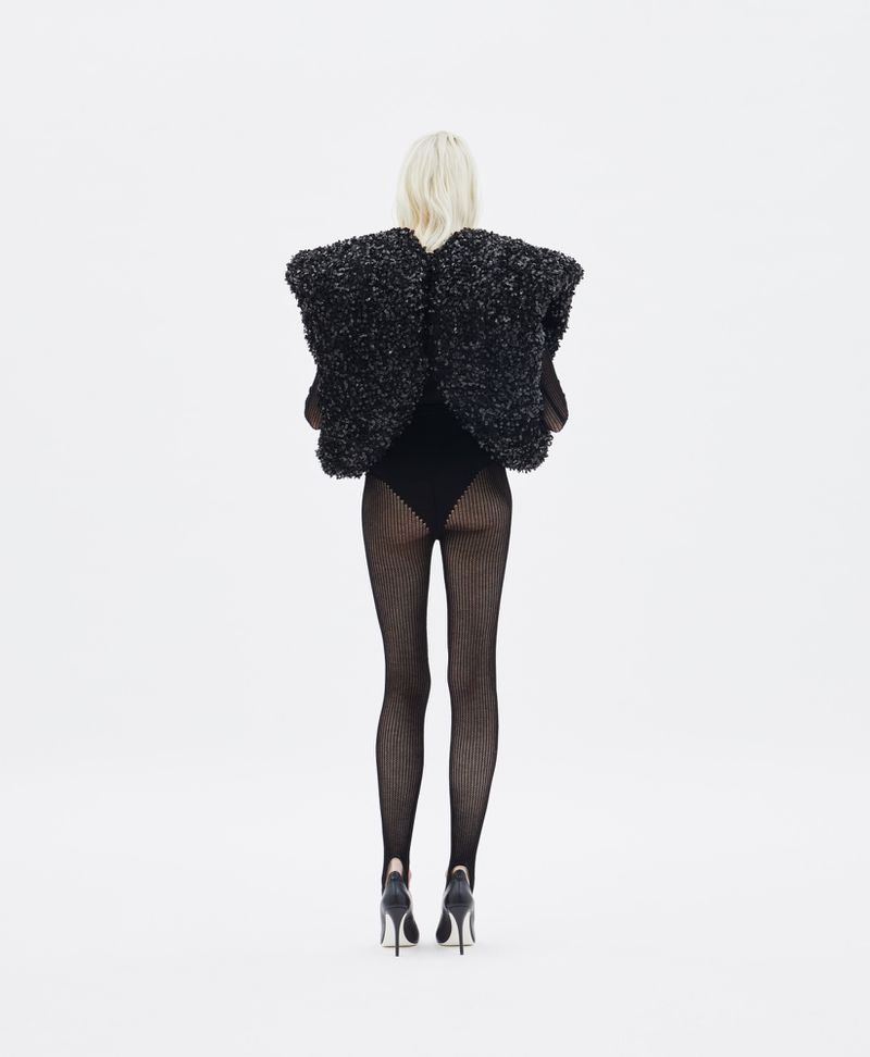 Woman wearing black fuzzy vest, bodysuit, textured tights, and black heels, standing against a white backdrop in a fashion shot.