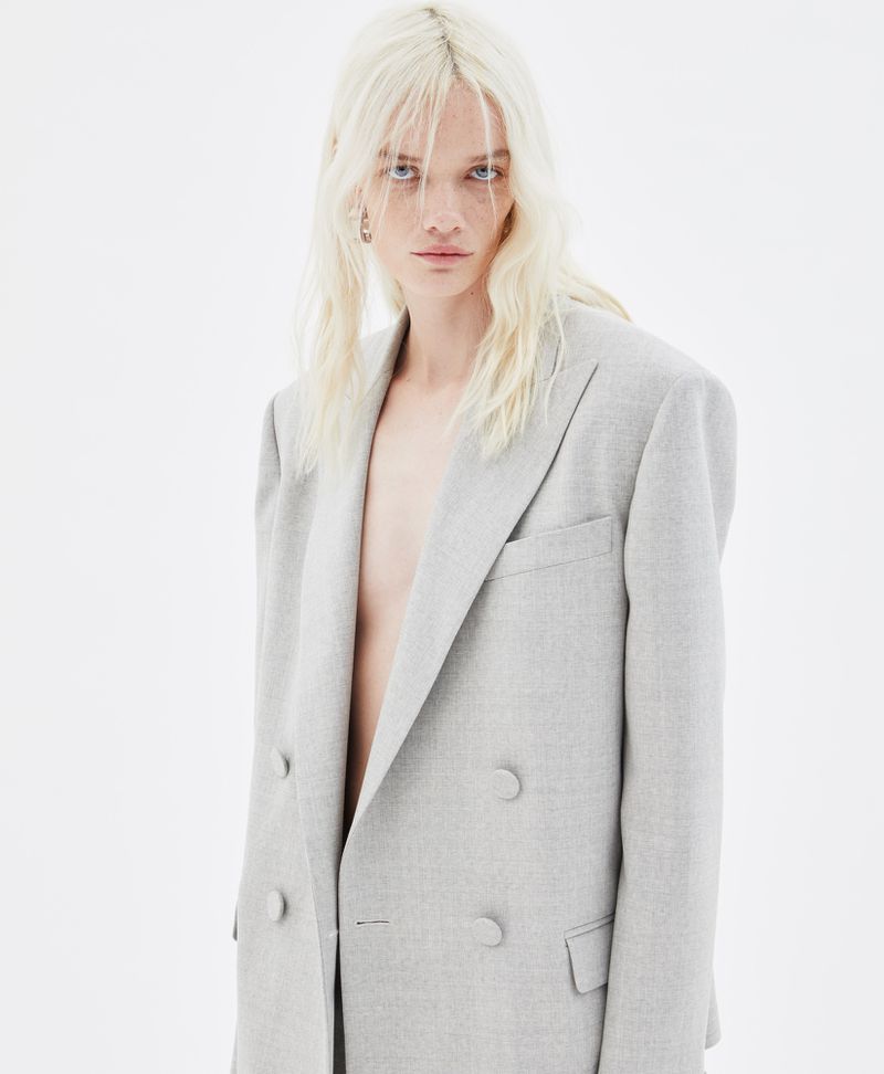 Model with blonde hair wearing a grey blazer against a plain white seamless studio background