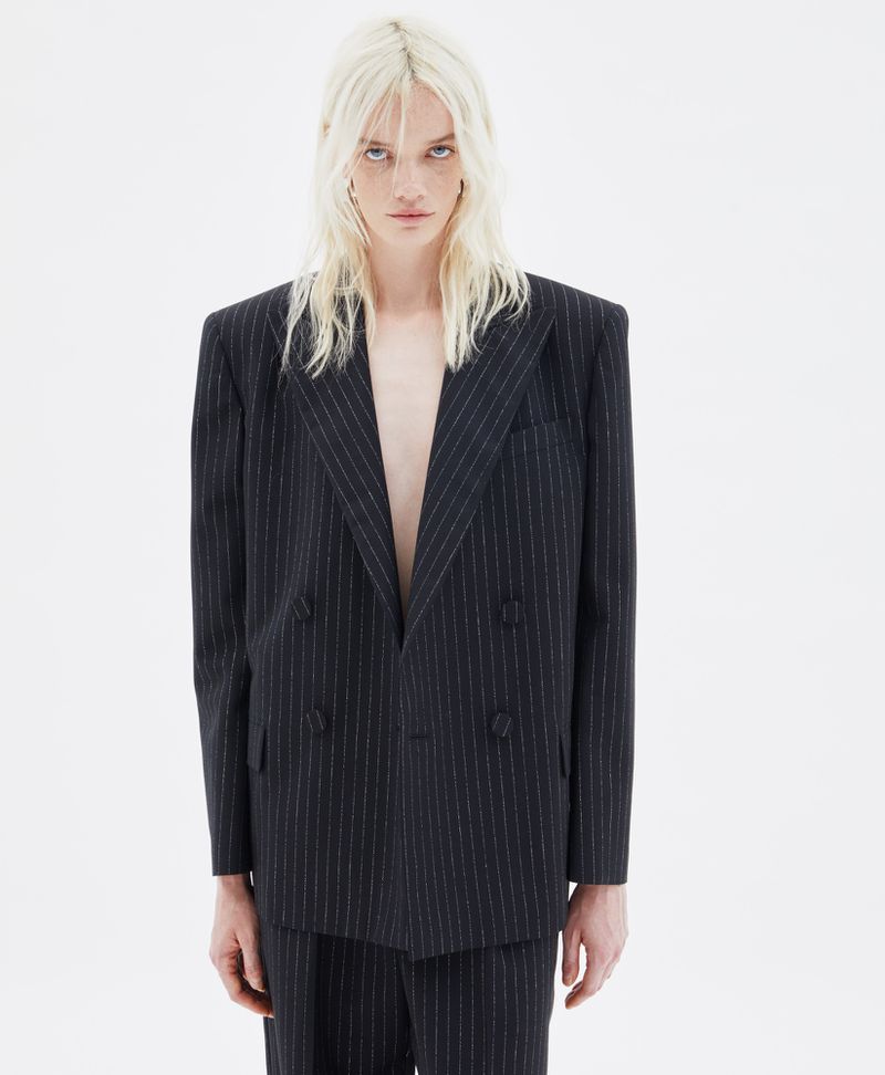 Portrait of a slender, androgynous person with white-blond hair wearing an oversized, dark pinstripe suit in a stark white setting.