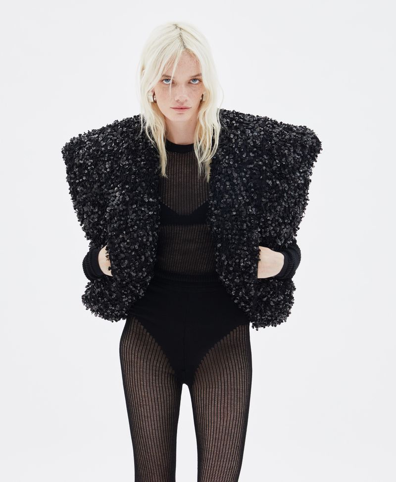 A woman with blonde hair wears a black sweater and unique fuzzy dark jacket in a fashion shot against a plain white backdrop.