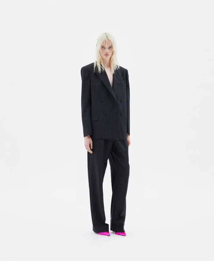 Model in a black pinstripe suit and bright pink shoes standing against a plain white background