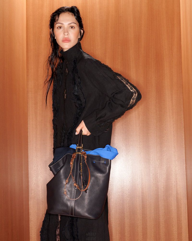 Dark-haired woman with a black leather tote bag poses indoors against a wood-paneled wall. She wears a black dress and has neutral expression.