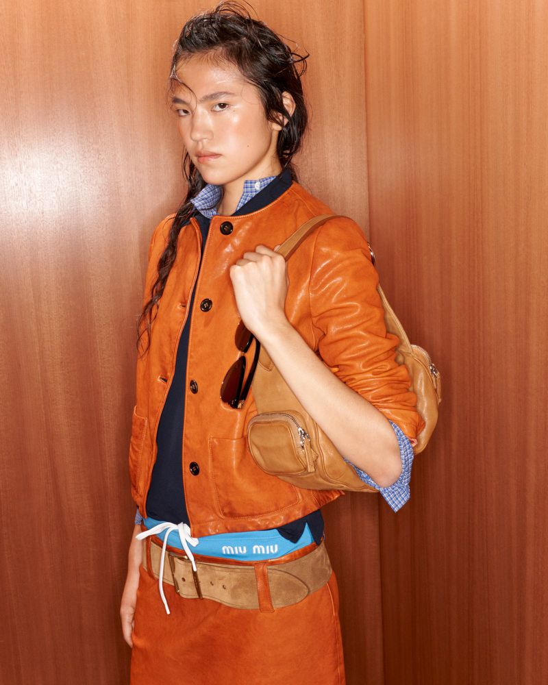 Model wearing orange leather jacket and matching skirt holding tan leather handbag against a wood paneled wall.