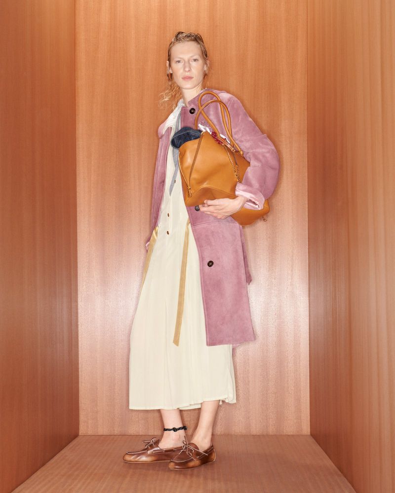 A woman in a rose coat and cream dress poses in a wooden corner holding a tan leather bag, exuding a fashion-forward vibe.