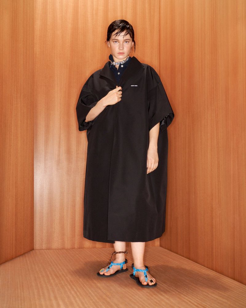 Woman in long black coat and teal sandals posing in corner with wood paneled walls and floor in a fashion editorial shot.