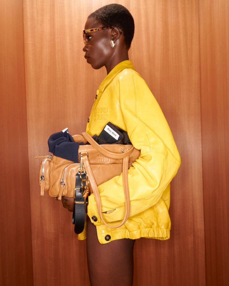 A stylish woman with short hair and glasses poses indoors wearing a yellow jacket, carrying a tan leather bag, against a brown panelled wall.