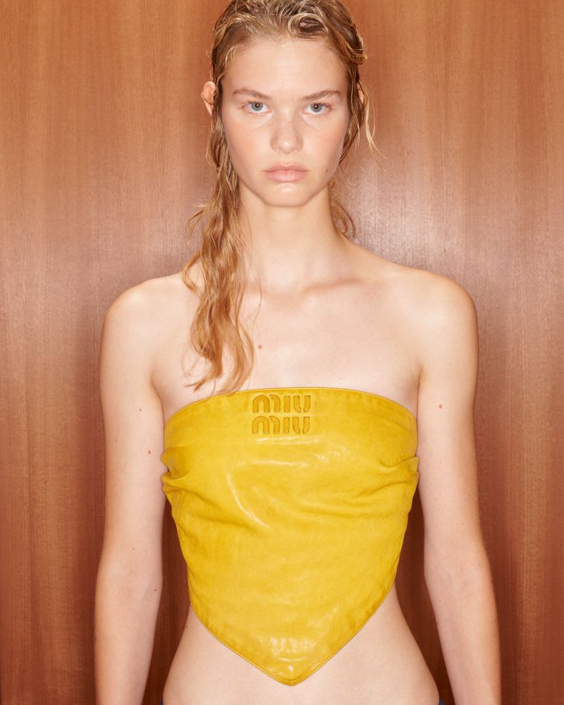 Close-up of a blonde woman wearing a yellow strapless top with logo against a wood background in a fashion-forward style.
