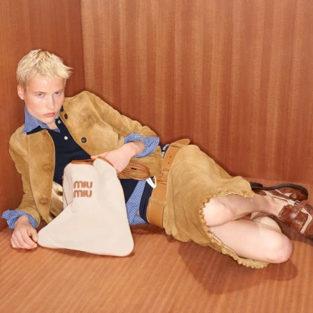 Model with blonde hair wearing tan suede jacket and skirt holding cream bag against wood paneled wall