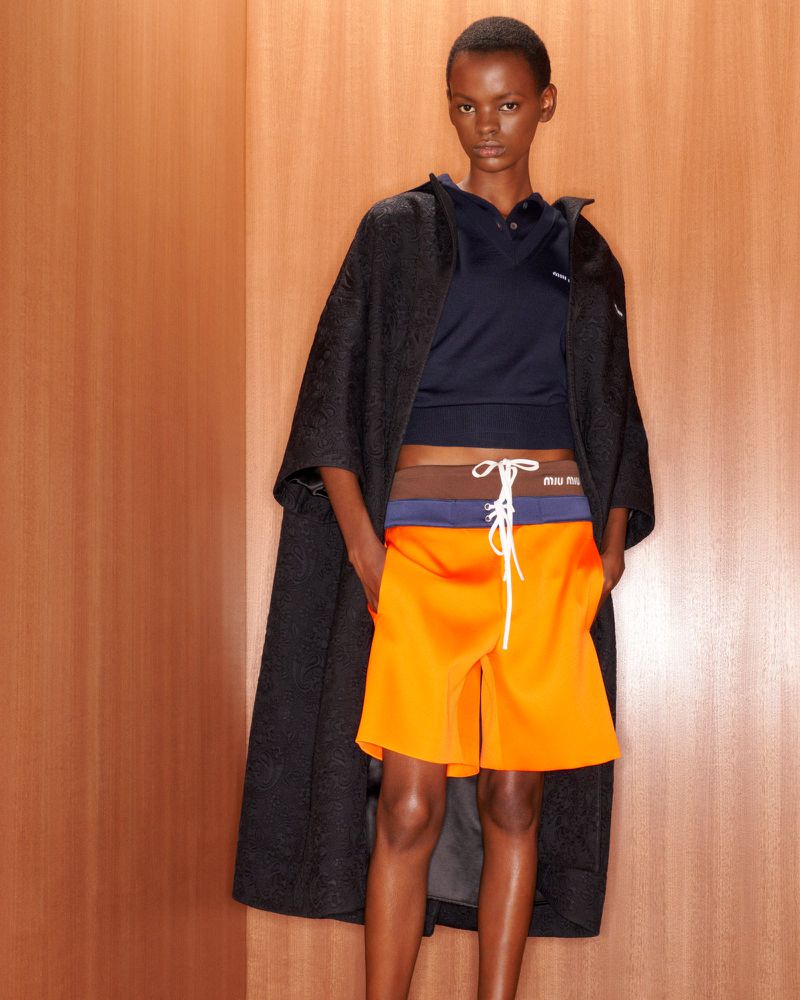 Fashion model poses in orange shorts, black coat and navy top against a peach-colored wall, showcasing a modern, high-fashion look.