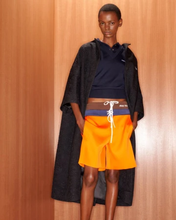 Fashion model poses in orange shorts, black coat and navy top against a peach-colored wall, showcasing a modern, high-fashion look.