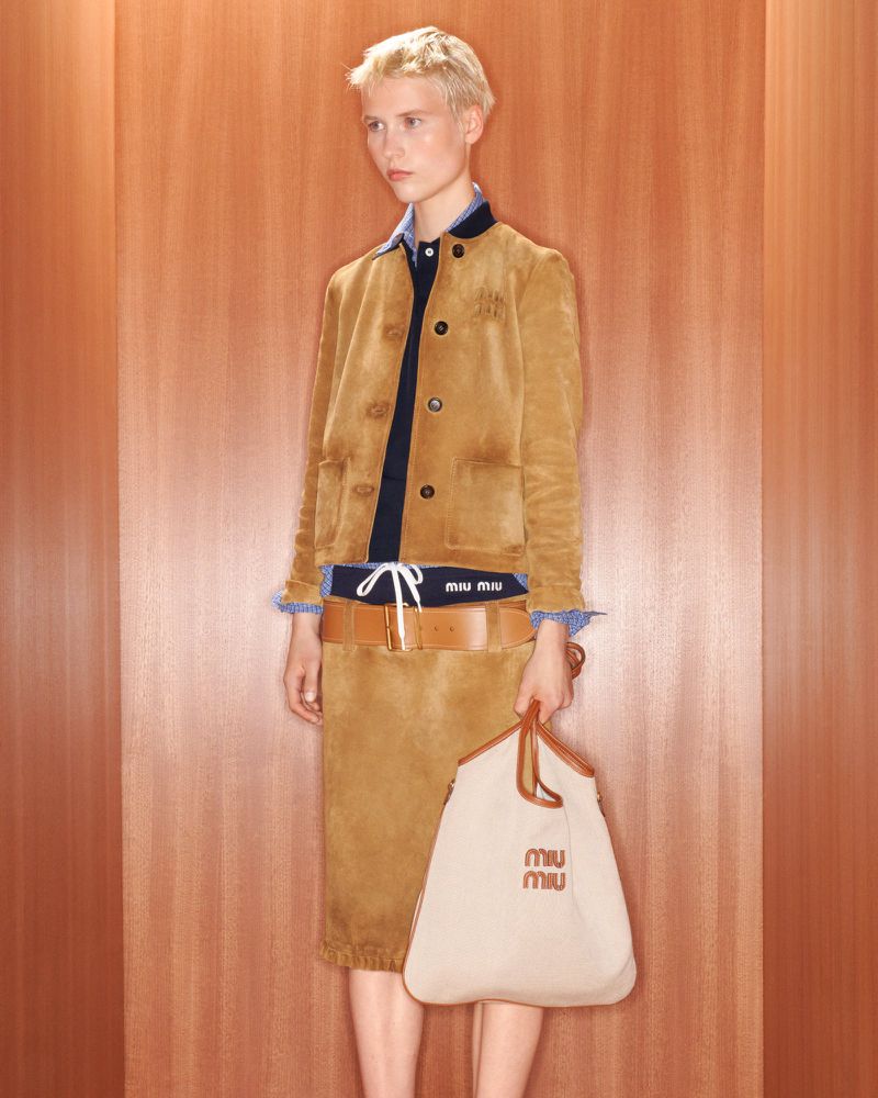 A young adult woman poses indoors in a brown suede skirt suit, navy shirt, and brown belt, holding a beige Miu Miu logo tote bag.