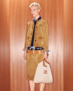 Model wearing a camel suede jacket and skirt holding a canvas shoulder bag against a wood panel wall