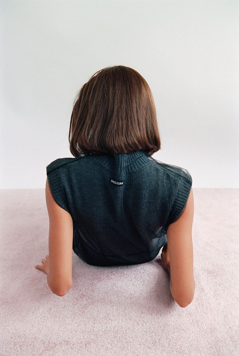 Back view of a person with short brown hair wearing a dark green knit vest, sitting on a pink carpet against a white wall.