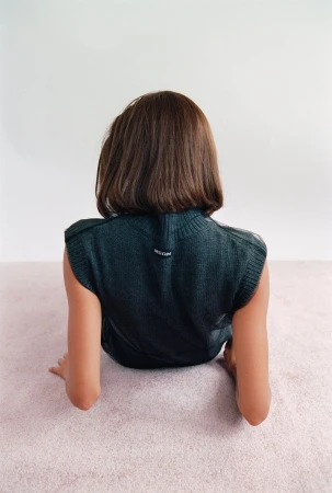 Back view of a person with short brown hair wearing a dark green knit vest, sitting on a pink carpet against a white wall.