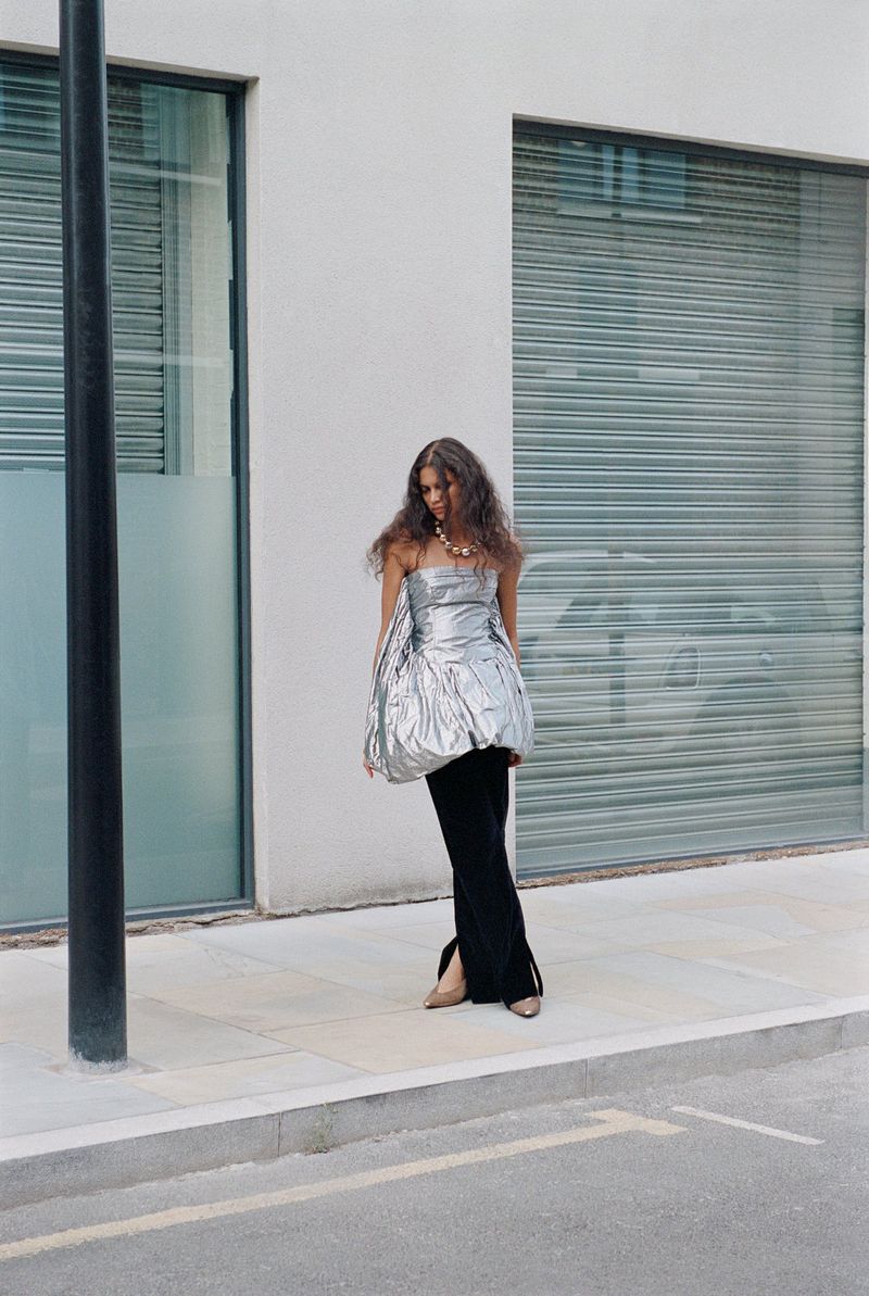 A model in a silver strapless dress and black pants poses outside a modern building, captured in a full shot, with street-level view.