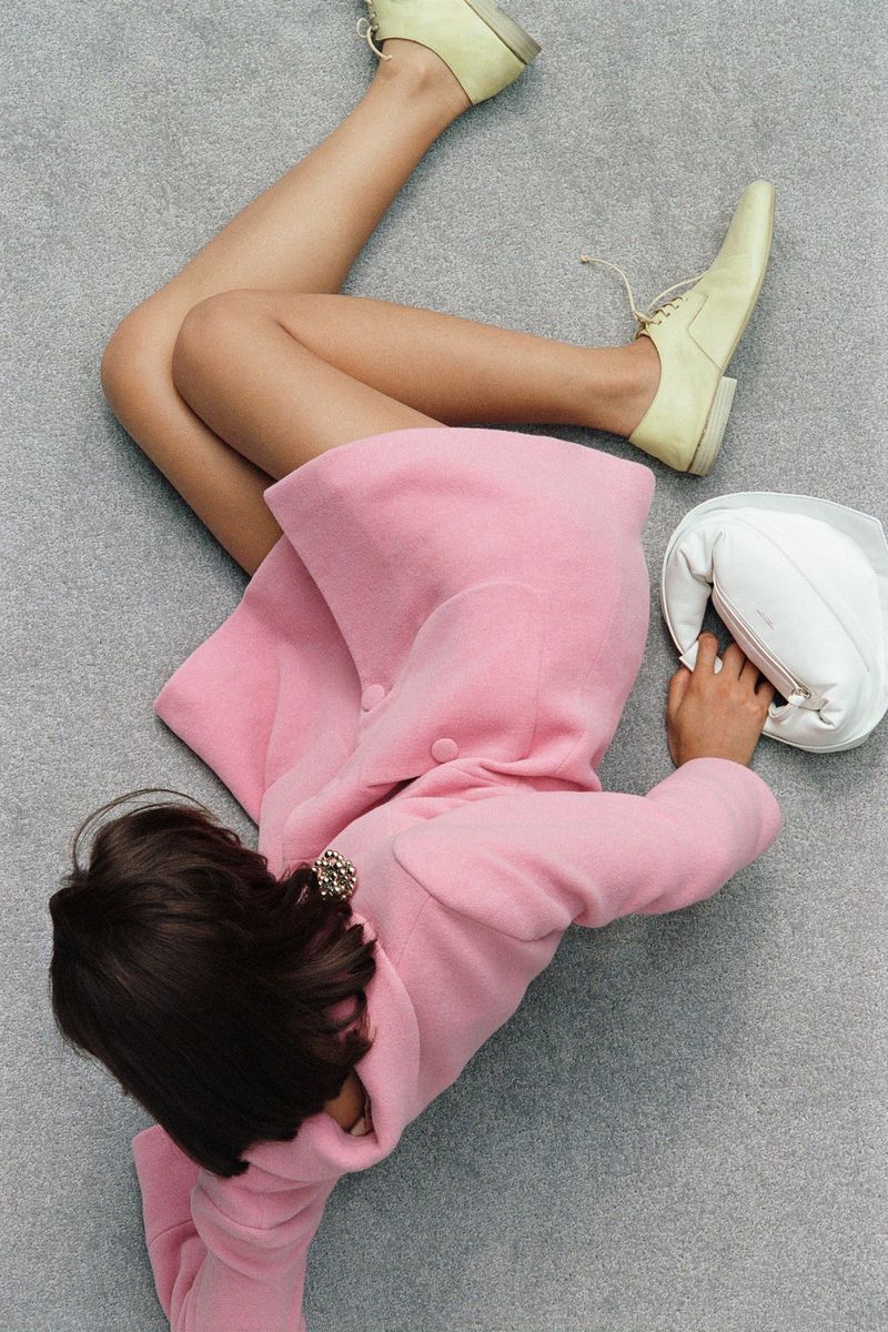 Overhead shot of a young woman in a pink coat lying on a gray carpet, wearing light yellow shoes and next to a white purse.