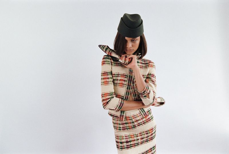 Fashionable woman in a plaid skirt suit and matching hat posing in a photo studio against a white backdrop. Dark olive hat.