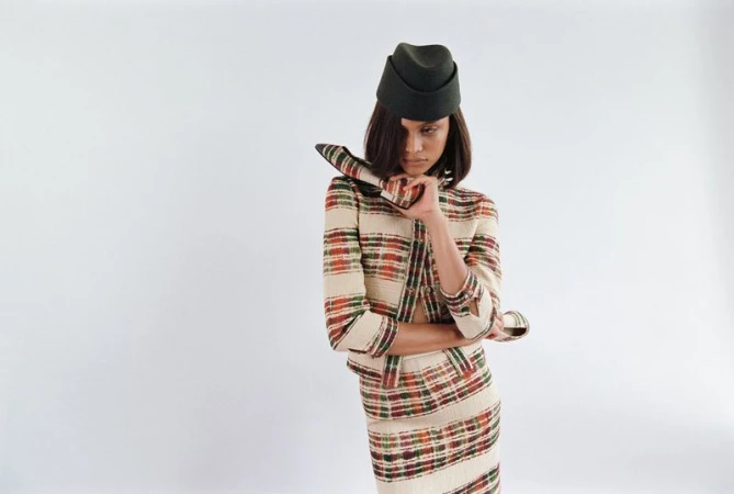 Fashionable woman in a plaid skirt suit and matching hat posing in a photo studio against a white backdrop. Dark olive hat.