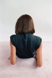 Model seen from behind with short brown hair wearing a dark sleeveless sweater, posed on a soft pink carpet.
