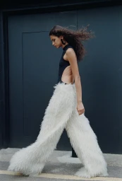 Model with long dark hair walking past a dark grey wall wearing a black bodysuit and shaggy white furry trousers.