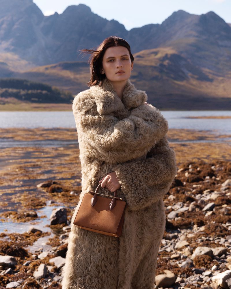 Adult woman in a shaggy coat poses with a brown handbag in a scenic outdoor environment featuring mountains and a lake.