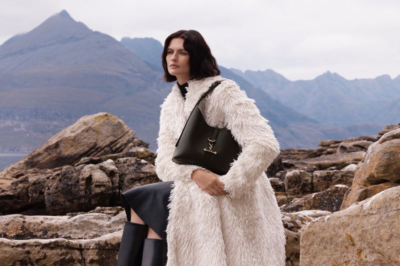 Fashionable woman poses with a handbag on a rocky terrain with a mountain range, wearing a white faux fur coat and black leather boots.