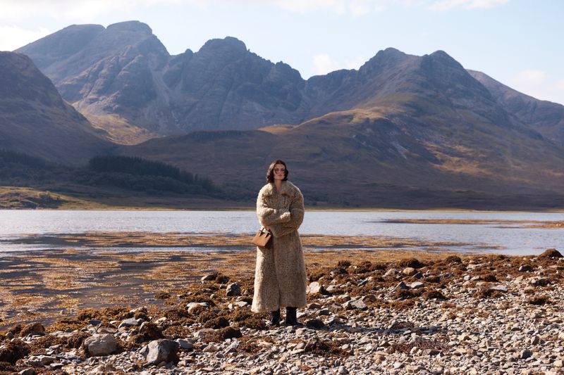 Adult woman in long coat standing on rocky shore with mountains, lake, and sky in the background on a sunny day in the outdoors.